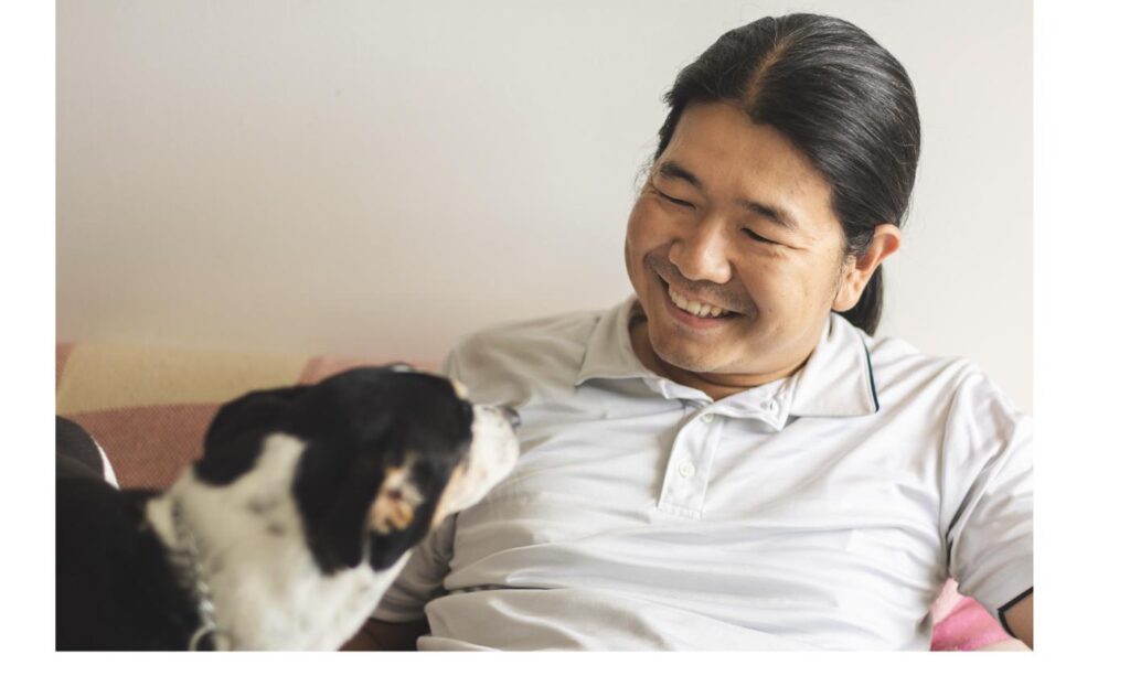 ソファで犬と笑顔で触れ合う男性、新しい家族との関係を一から築く様子をイメージ
